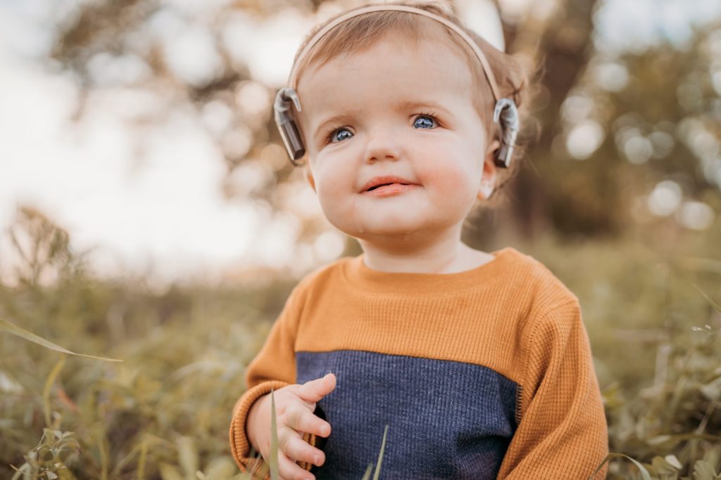 A toddler with a thoughtful expression, wearing a cute brown and navy pullover, enjoys the outdoors with tiny headphones on. This scene is beautifully captured in a children's book about raising a deaf child,