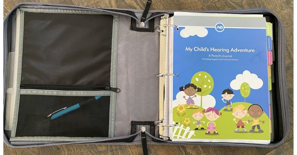 A binder opened on a wooden surface, displaying a learning resource titled "My Child's Hearing Adventure: A Parent's Journal" on the right, and a compartment with a pen on the left side.