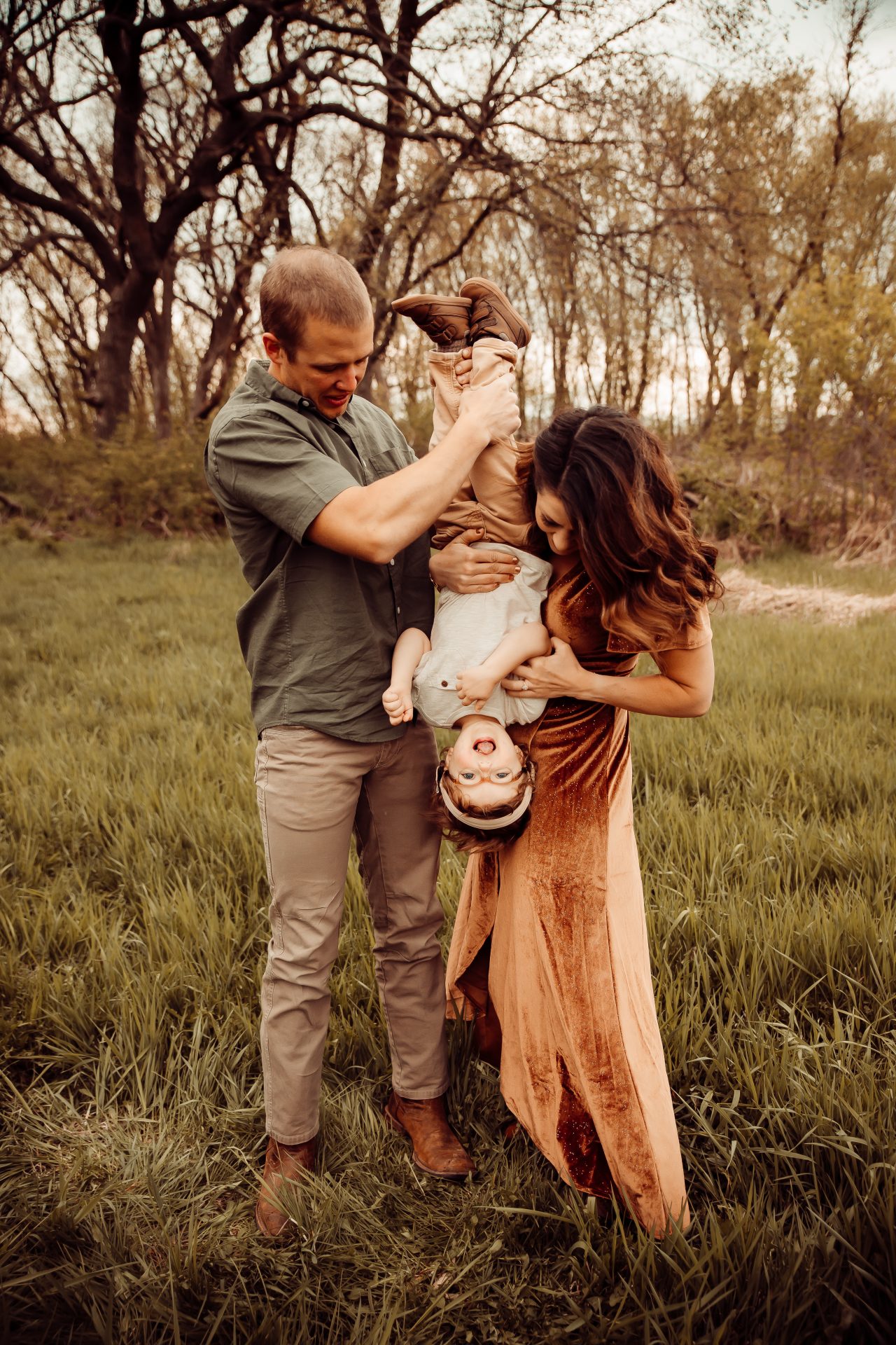 A joyful moment in a grassy field as a couple plays with their little one, raising a deaf child with love and inclusion, holding the child upside down, with laughter in the air.