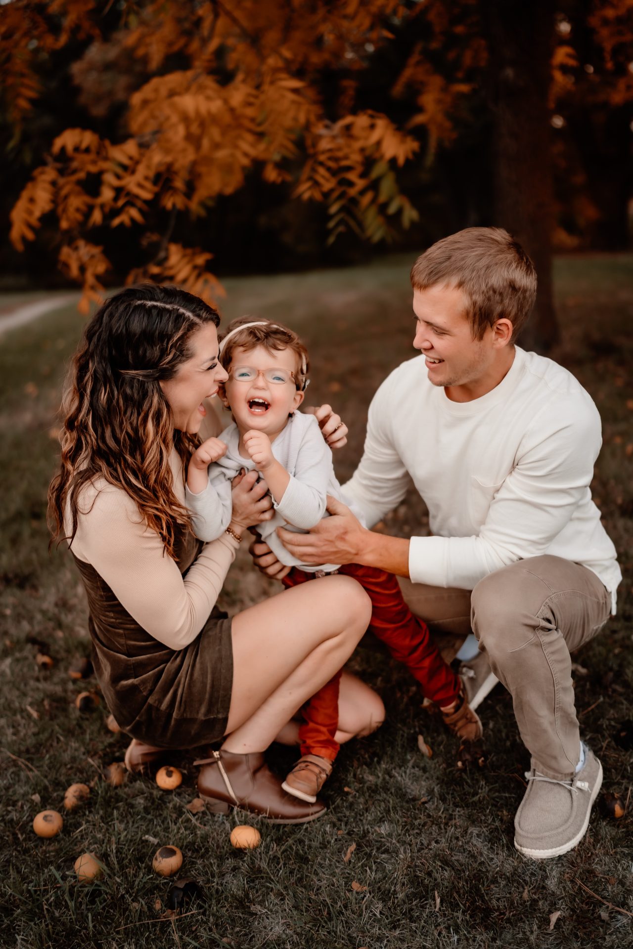 Brandon, Beth and Cooper kneel in grass in front of a colorful fall tree. Beth is an inclusion advocate with a focus on raising a deaf child.