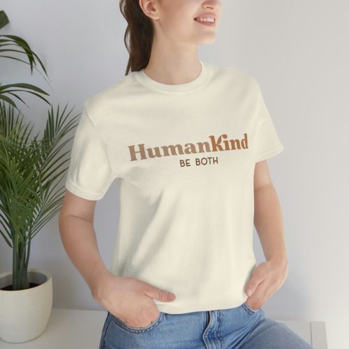 Woman smiling and posing in a casual setting, wearing a beige t-shirt with the 'HumanKind Be Both' message printed on the front, embodying the spirit of inclusion.
