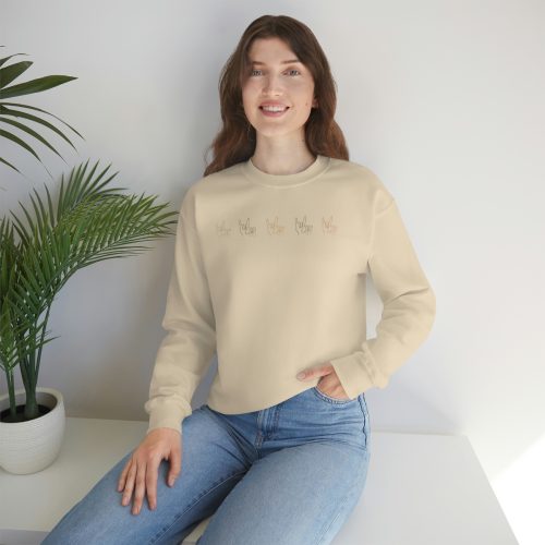 A smiling woman in a casual 'My Love' crewneck sweatshirt and blue jeans sitting against a white wall, with a potted plant beside her, reads a children's book about disabilities.