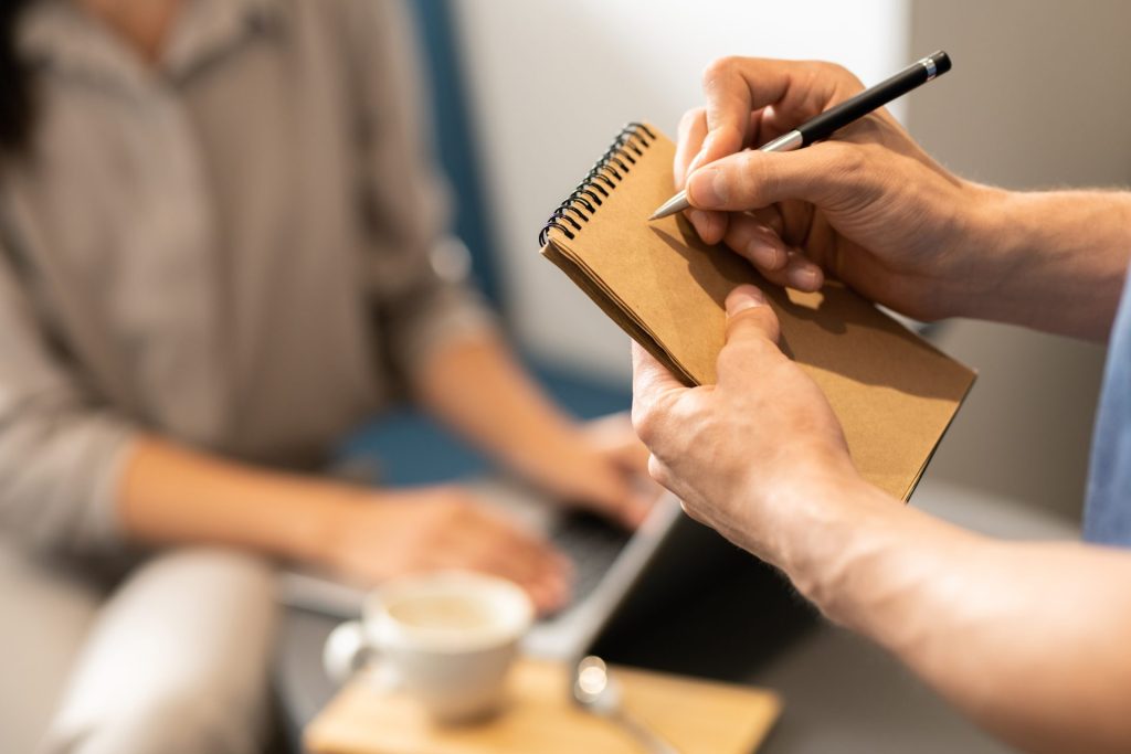 Close-up of two people in a meeting; one is holding a notebook and pen, while the other has a laptop beside a coffee cup, emphasizing a collaborative work environment.