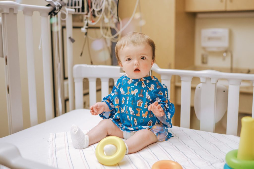A baby with light hair sits on a hospital bed wearing a blue gown with cartoon animals. The baby looks toward the camera with a curious expression. In front of the baby are colorful rings and a striped blanket, with medical equipment visible in the background.